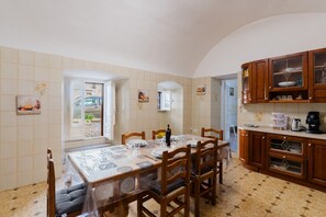 Un'area pranzo con tavolo apparecchiato per quattro persone, sedie di legno, una cucina con pensili e un pavimento piastrellato.