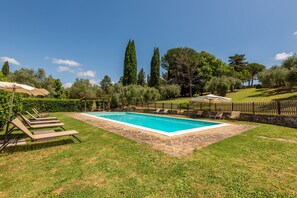 A rectangular swimming pool with a stone border, surrounded by a grassy lawn, and shaded by umbrellas.
