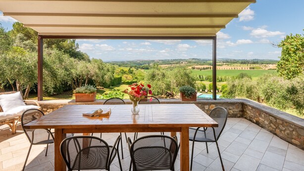 Une terrasse extérieure couverte, avec une table et des chaises en bois, un vase de fleurs et une vue sur la campagne.
