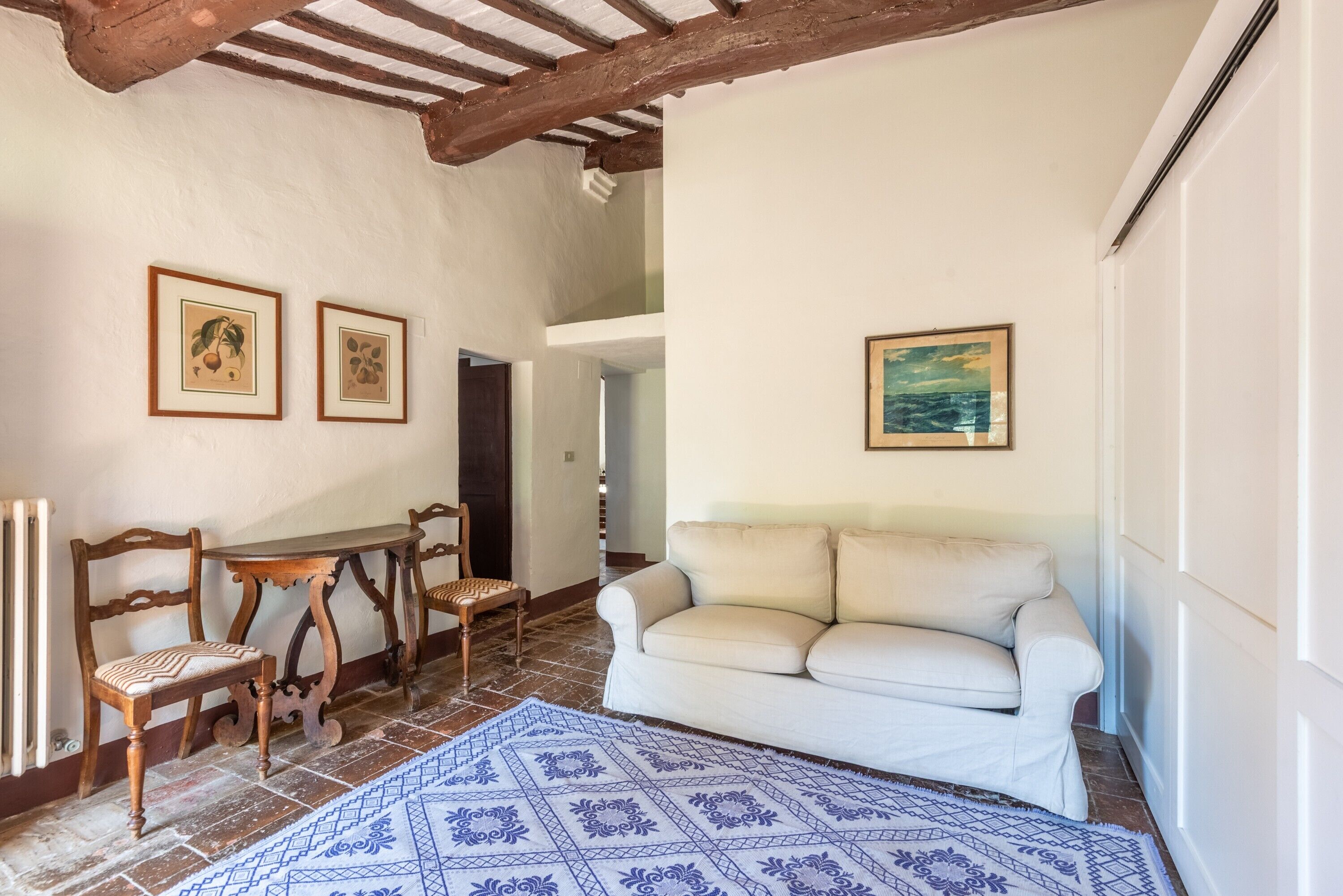 A room with a white sofa, wooden chairs, a small table, and a patterned rug.