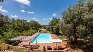 Ein rechteckiger Swimmingpool mit überdachtem Bereich, umgeben von einer Ziegelterrasse und Blick auf eine grüne Landschaft.