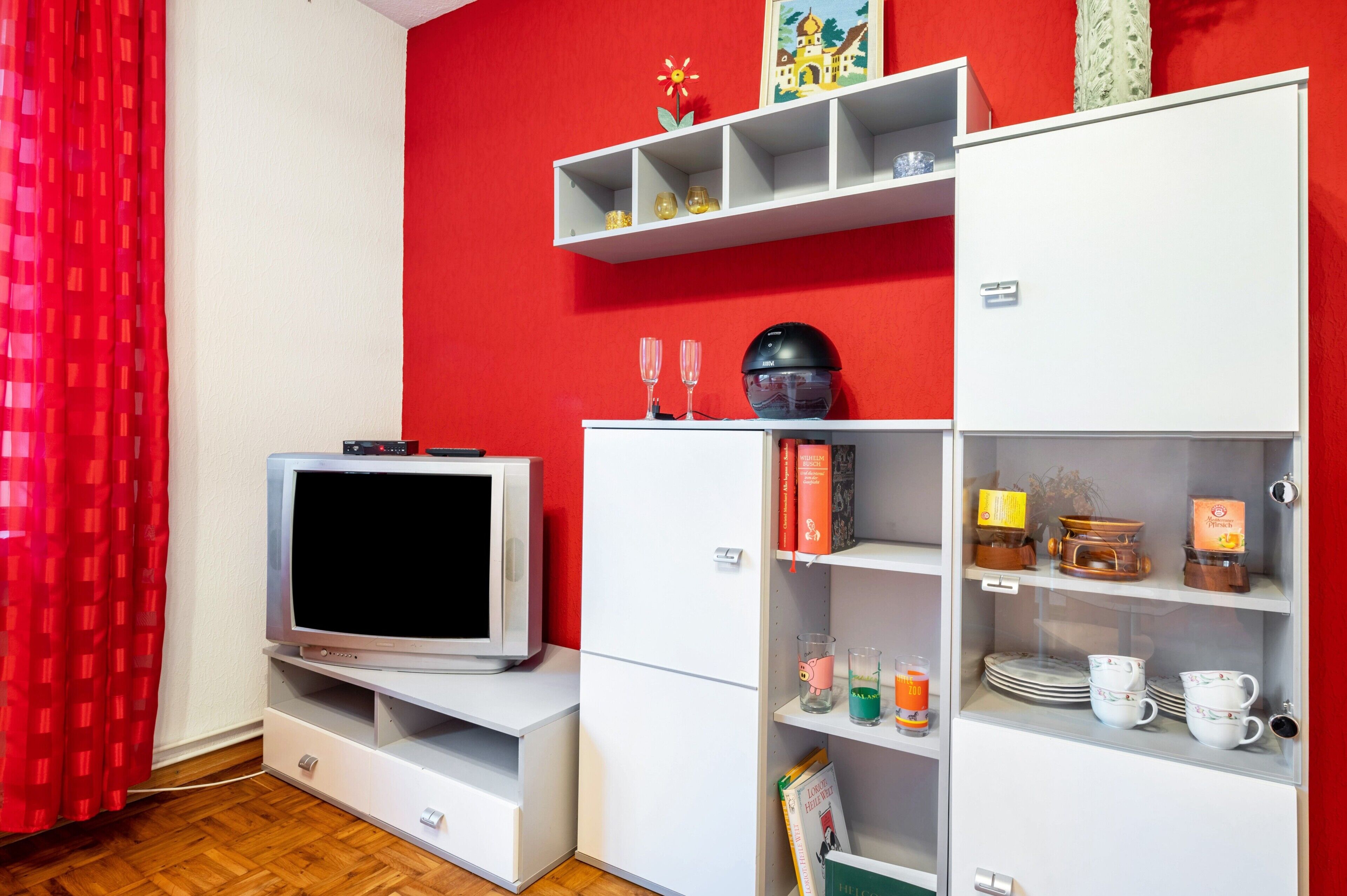 A room with a television on a stand, a white cabinet with shelves, and a red wall.