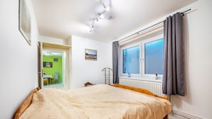 Een slaapkamer met een bed, een raam met gordijnen, een radiator en een deur die naar een andere kamer leidt.