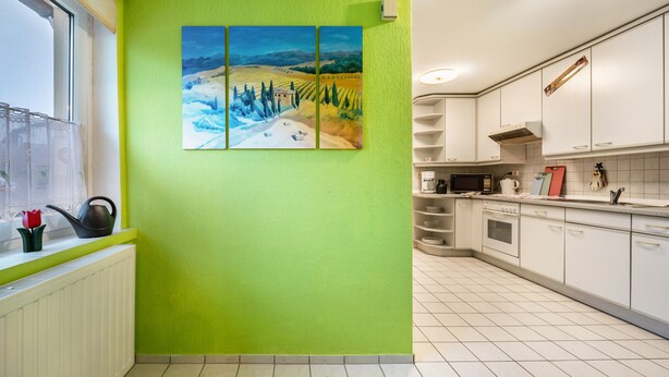 Een keuken met witte kasten, een raam met uitzicht en een groene muur met een schilderij.