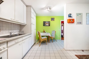 Une cuisine avec des meubles blancs, une table à manger avec deux chaises, un mur vert et une petite table sur laquelle est posé un vase.