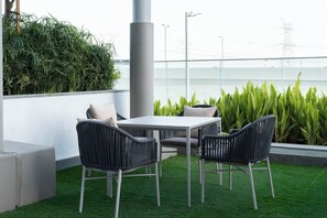 Um terraço no telhado com uma mesa branca e duas cadeiras de vime, rodeado por plantas verdes e uma vista desimpedida da linha do horizonte da cidade.