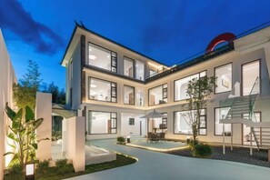 Una casa moderna de varios pisos con amplios ventanales, un espacio cubierto para sentarse al aire libre y un camino de entrada bien iluminado.