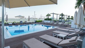 A rooftop pool area with lounge chairs and umbrellas, surrounded by greenery and a cityscape view. - Modern Studio in Azizi Riviera 46  (Dubai)