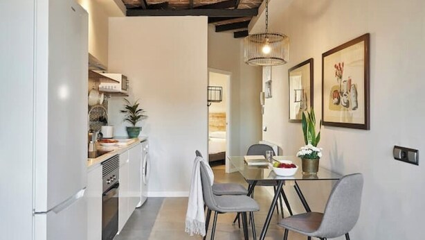A modern kitchen with a white refrigerator, stainless steel appliances, and a wooden ceiling. There is a dining area with a glass table and grey chairs, and a hallway leading to a bedroom. - 1-BR flat with terrace -Eixample (Barcelona)