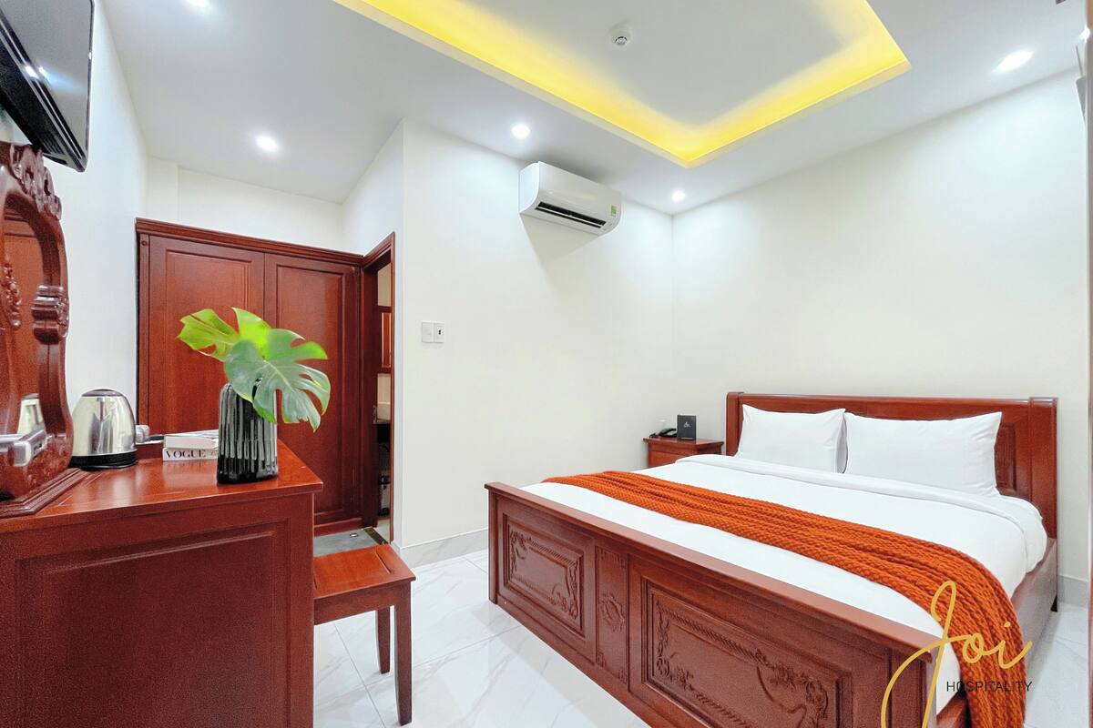 A hotel room with a wooden bed, a desk with a plant, a chair, and a nightstand.