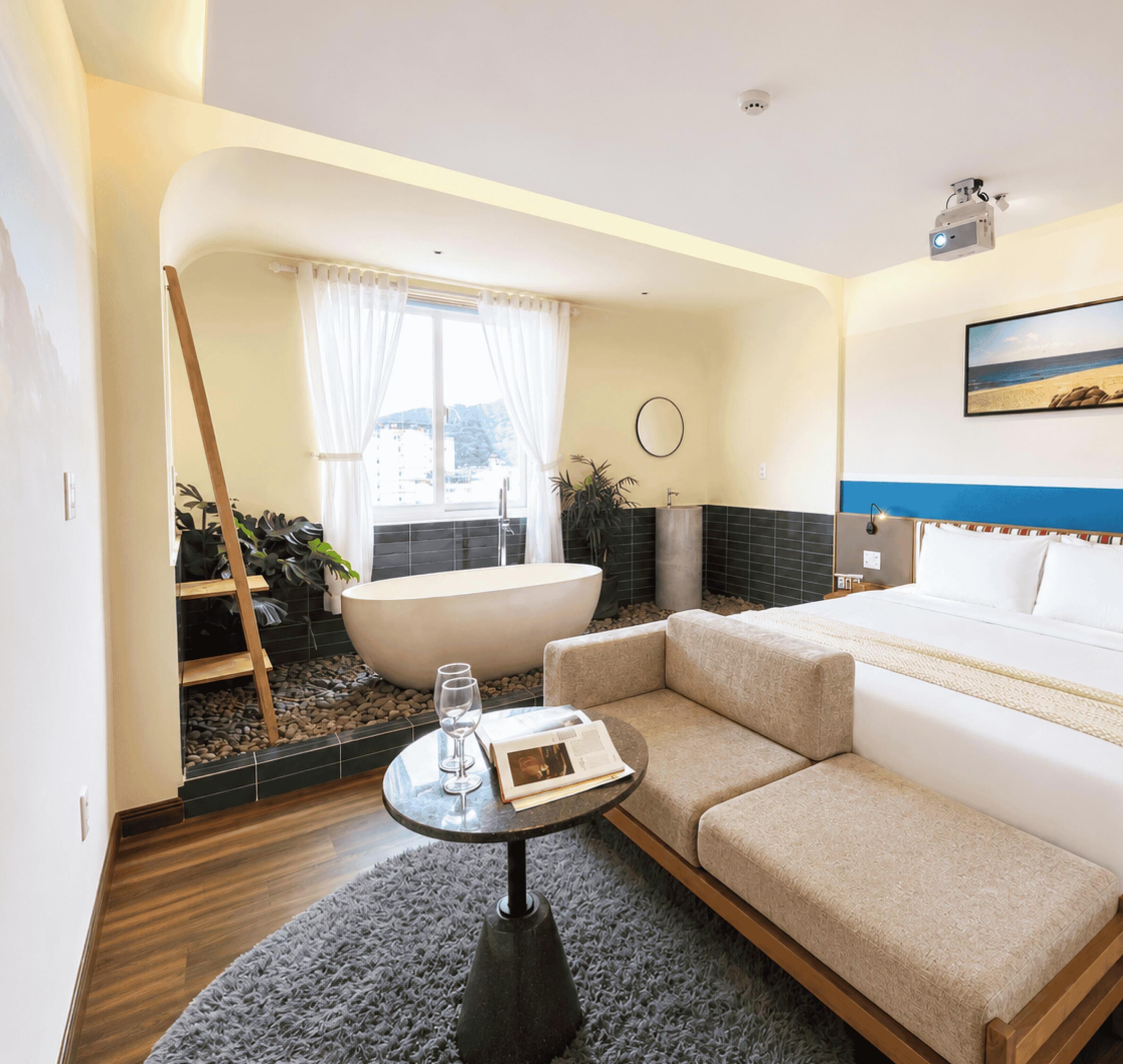 A modern hotel room with a sofa, a small table with glasses, a bathtub, and a view of the mountains.
