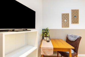 A modern living room with a flat-screen TV on a white entertainment unit, a wooden dining table with a bench, a potted plant, and two decorative wall panels.