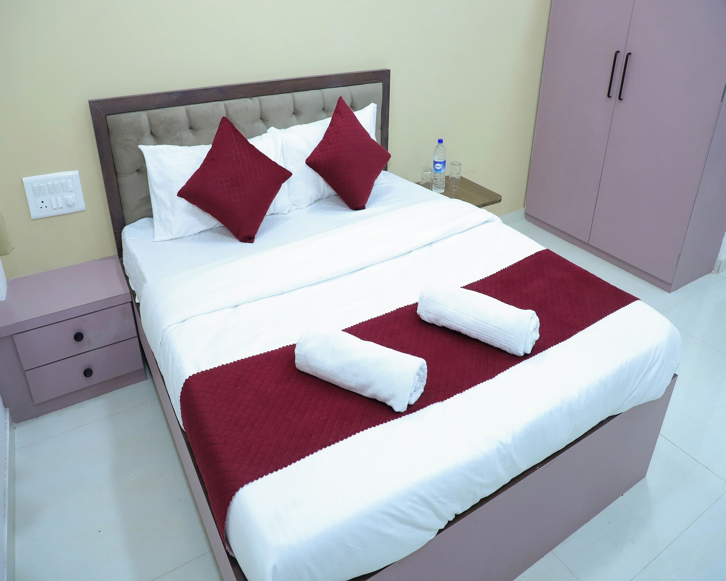 A neatly made bed with white and red bedding, a water bottle, and glasses on a nightstand.