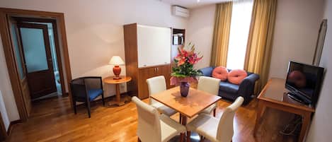 A modern hotel room with a wooden floor, a dining table with four chairs, a sofa, a TV, and a small side table with a lamp.