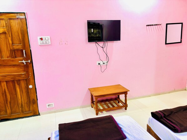 A room with a wooden door, a television mounted on the wall, a small wooden table, and two beds with blankets. - Tirupati Yatri Nivas (Aundha Nagnath)
