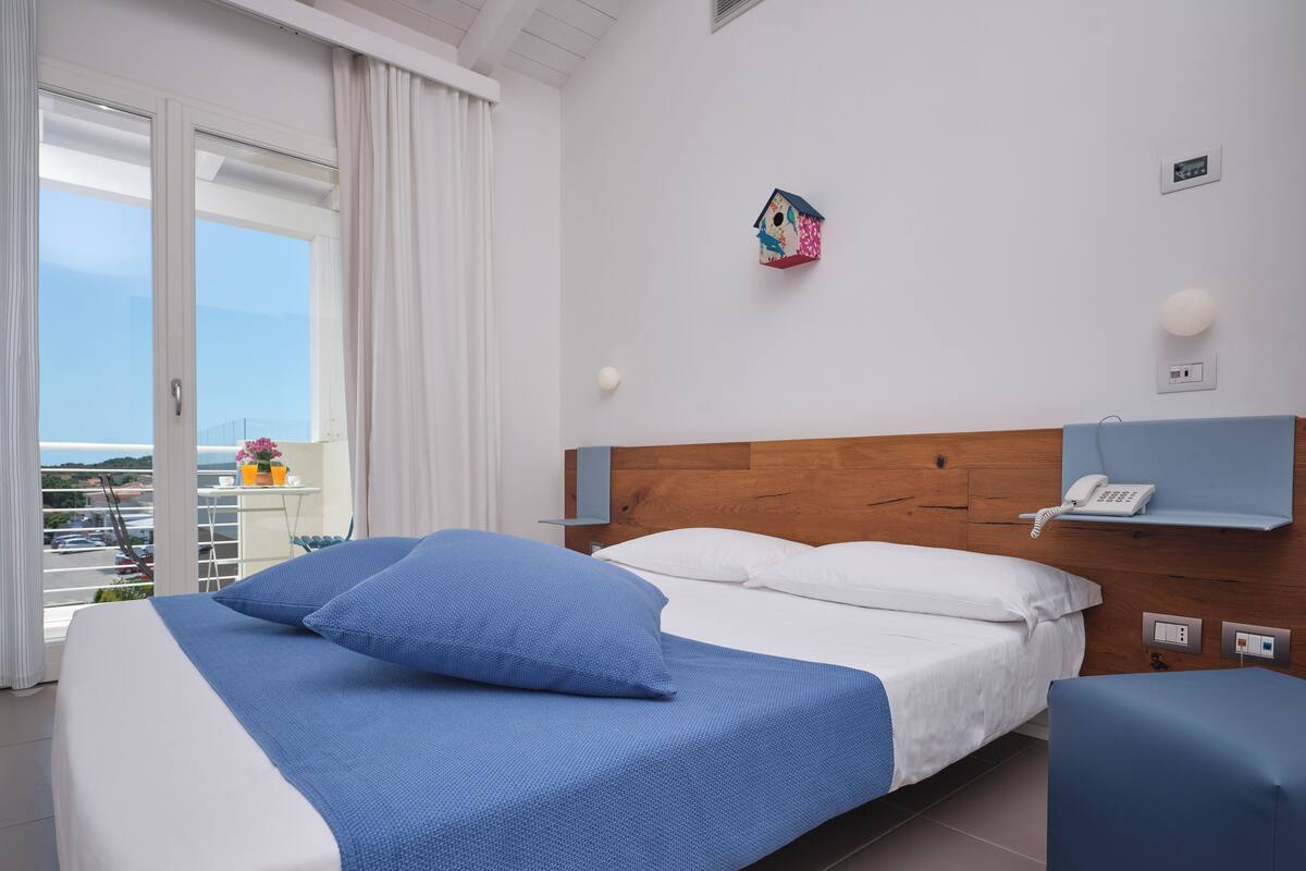Habitación de hotel con cama, mesita de noche, teléfono, luz empotrada en la pared y vistas al exterior.