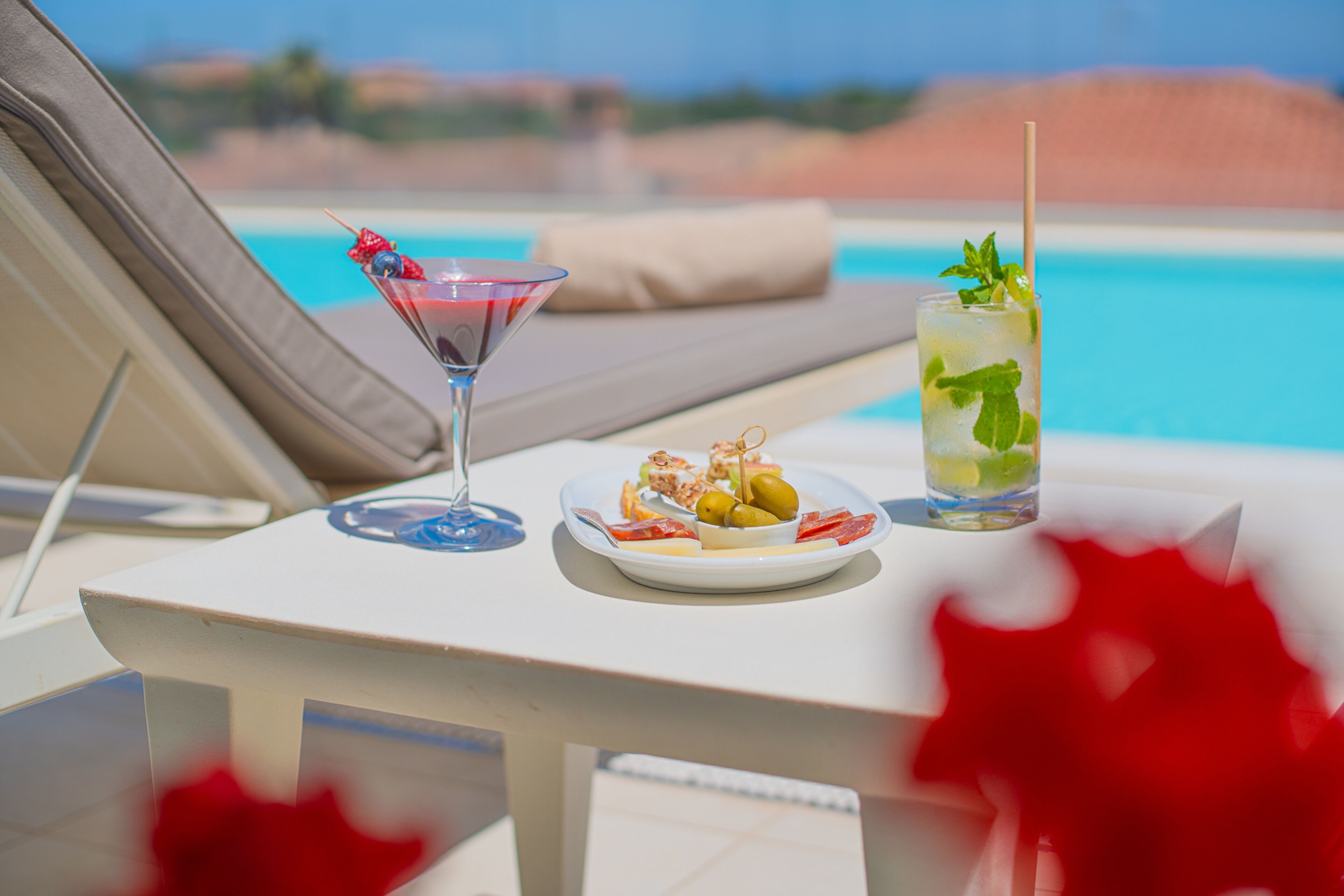 Une table au bord de la piscine avec un verre à martini, un grand verre avec une paille et une assiette de nourriture.