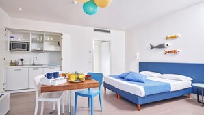 Una habitación de hotel moderna con espacio para cocinar, una mesa de comedor con sillas, una cama con ropa de cama azul y peces decorativos en la pared.