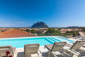 A swimming pool with lounge chairs, a mountain in the background, and a coastal town. - Hotel with panoramic swimming pool in porto San Paolo (Porto San Paolo)