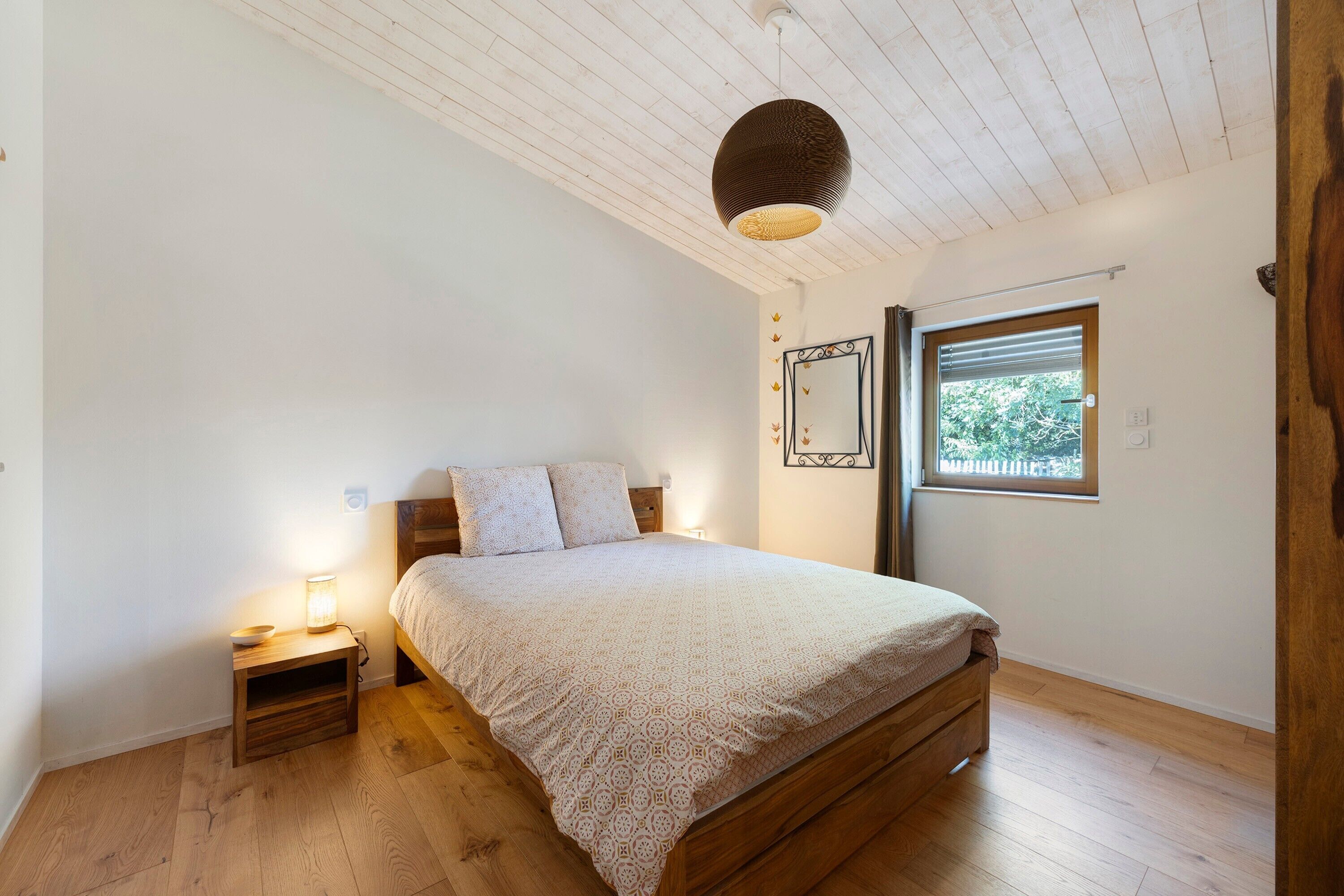 Et soveværelse med en træseng, et natbord med en lampe, et vindue med persienner og en loftlampe.