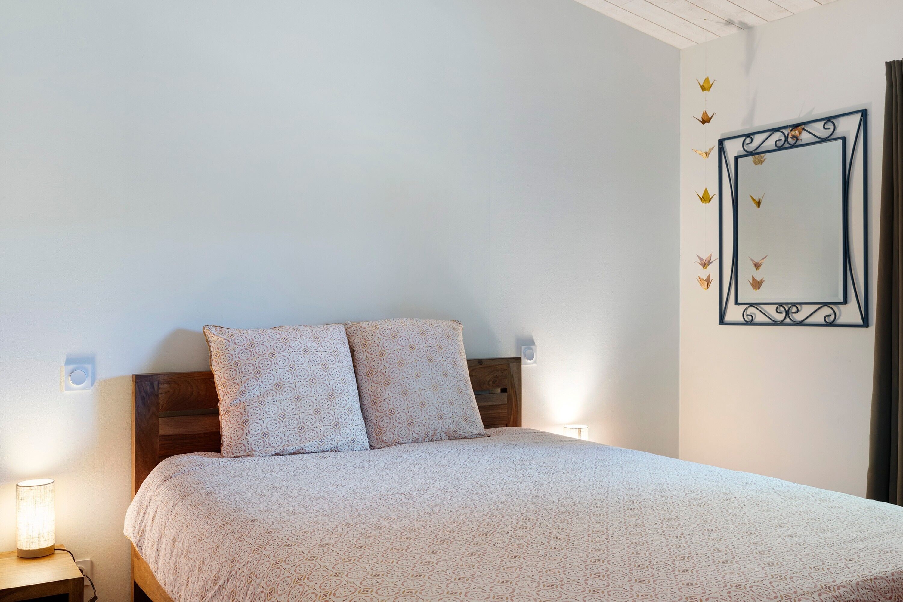 A bedroom with a bed, two patterned pillows, a bedside lamp, a wall-mounted mirror, and a decorative wall piece.