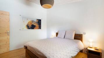Een slaapkamer met een houten bed, een nachtkastje met een lamp, een aan de muur hangend abstract schilderij en een houten deur.