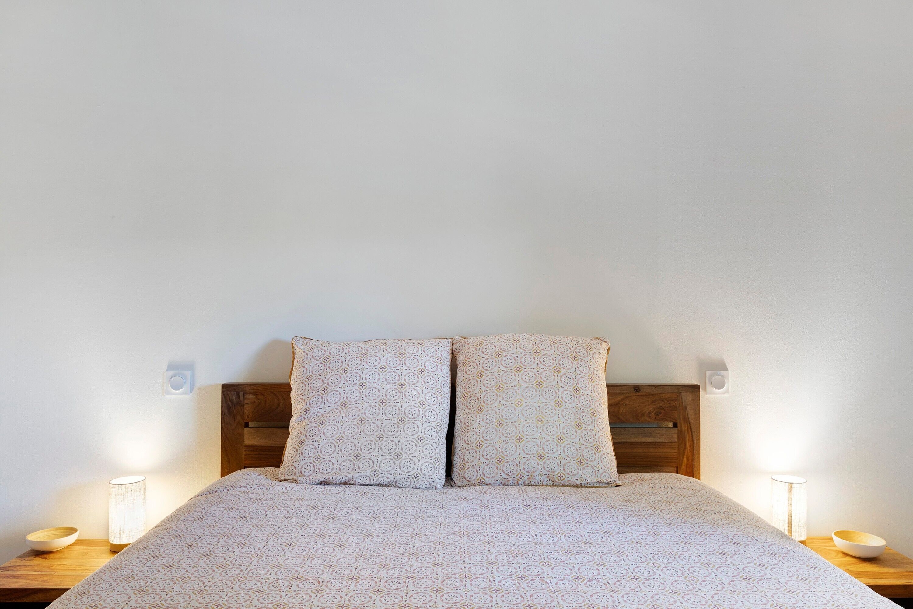 Una cama con colcha estampada, dos almohadas, cabecera de madera, lámparas de noche y una mesita de noche con un cuenco.