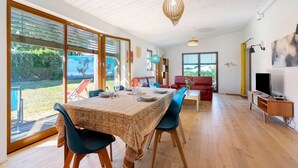 Un comedor amplio con una mesa dispuesta para cuatro personas, ventanas grandes, un sofá rojo y un televisor sobre un mueble de madera.