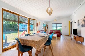 Un'ampia sala da pranzo con un tavolo apparecchiato per quattro persone, grandi porte scorrevoli in vetro che si aprono su un giardino, un divano rosso e un televisore su un supporto in legno.