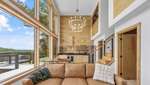 A spacious living area with a leather sofa, a chandelier, and large windows offering a view of the outdoors. - Elevated Invisible Cabin, Hot Tub, Views (Marshall)