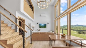 A spacious living area with a wooden staircase, a large window offering a view of the countryside, and a modern chandelier. - Elevated Invisible Cabin, Hot Tub, Views (Marshall)