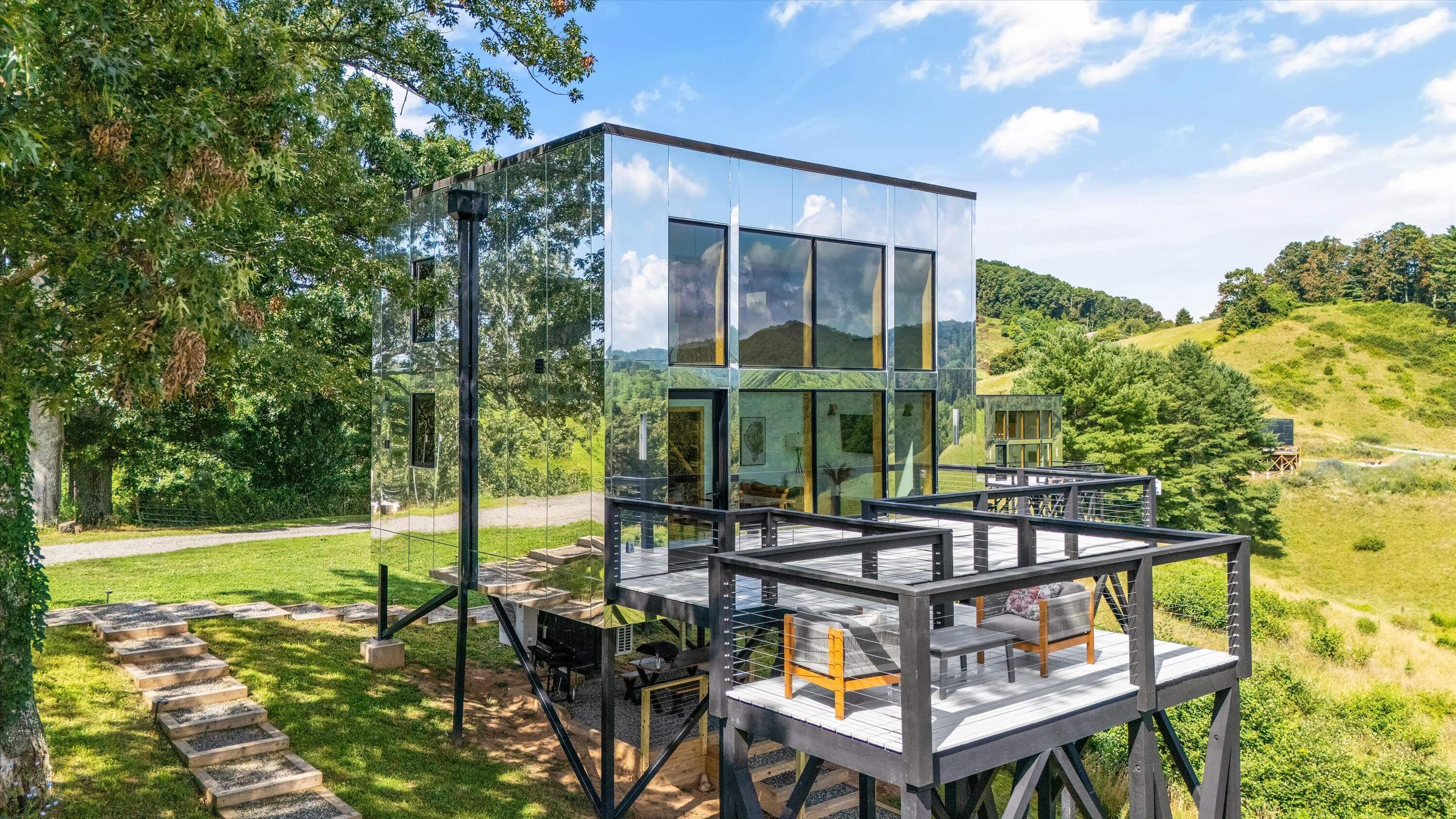 Une maison moderne en verre, dotée d’une terrasse et d’un espace salon extérieur, entourée d’une végétation luxuriante et de collines.