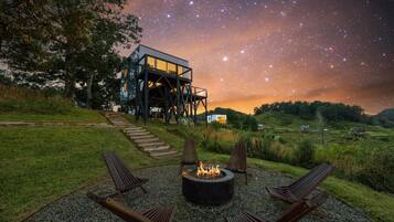 Un ciel étoilé, un foyer avec des chaises autour, et une maison moderne dotée d’un balcon en verre.