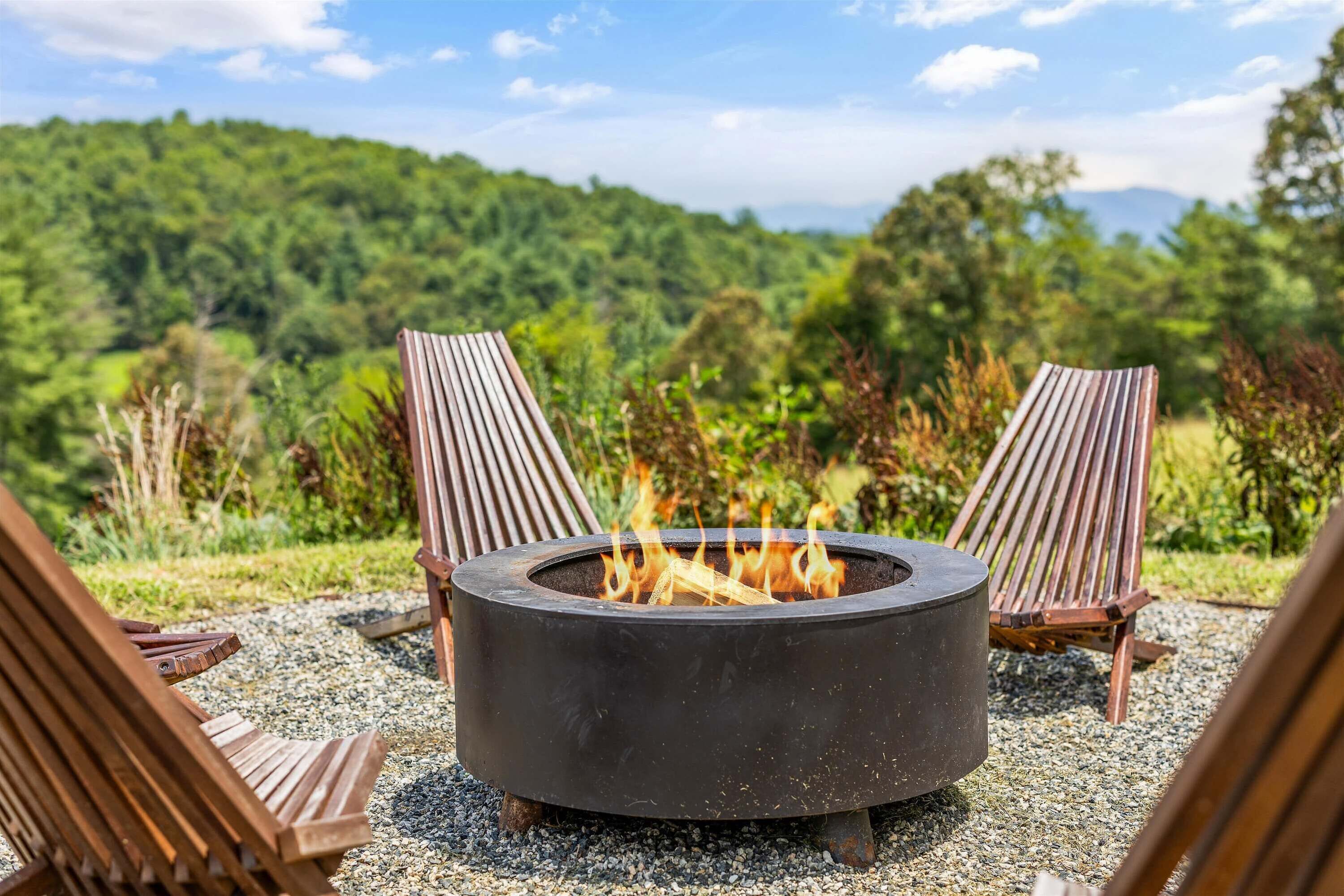 A circular outdoor fire pit with two wooden chairs facing it, surrounded by gravel, set against a backdrop of lush greenery and a clear sky.