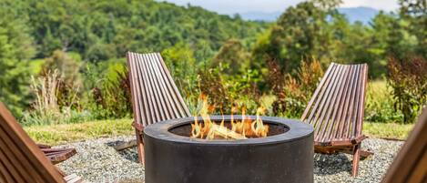 A circular outdoor fire pit with two wooden chairs facing it, surrounded by gravel, set against a backdrop of lush greenery and a clear sky.