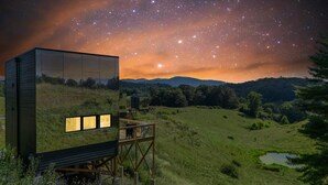 Eine moderne Glasarchitektur mit zahlreichen Fenstern, einem Holzboden und Blick auf einen sternenübersäten Nachthimmel über einer Hügellandschaft mit Gras.