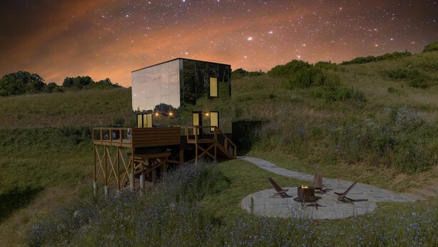 Eine moderne, zweistöckige Holzhütte mit Glasfassade, gelegen auf einem grasbewachsen Hügel unter einem Sternenhimmel.
