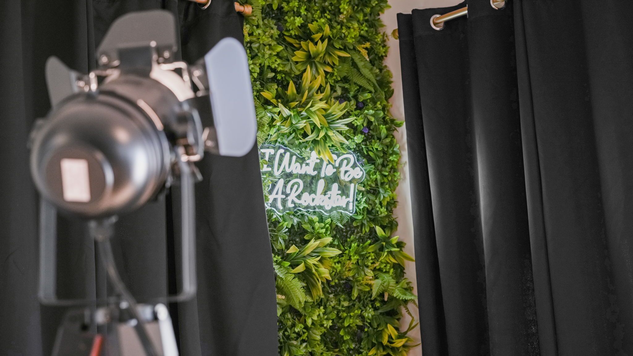 A camera on a tripod, a black curtain, and a wall with green foliage and a neon sign.