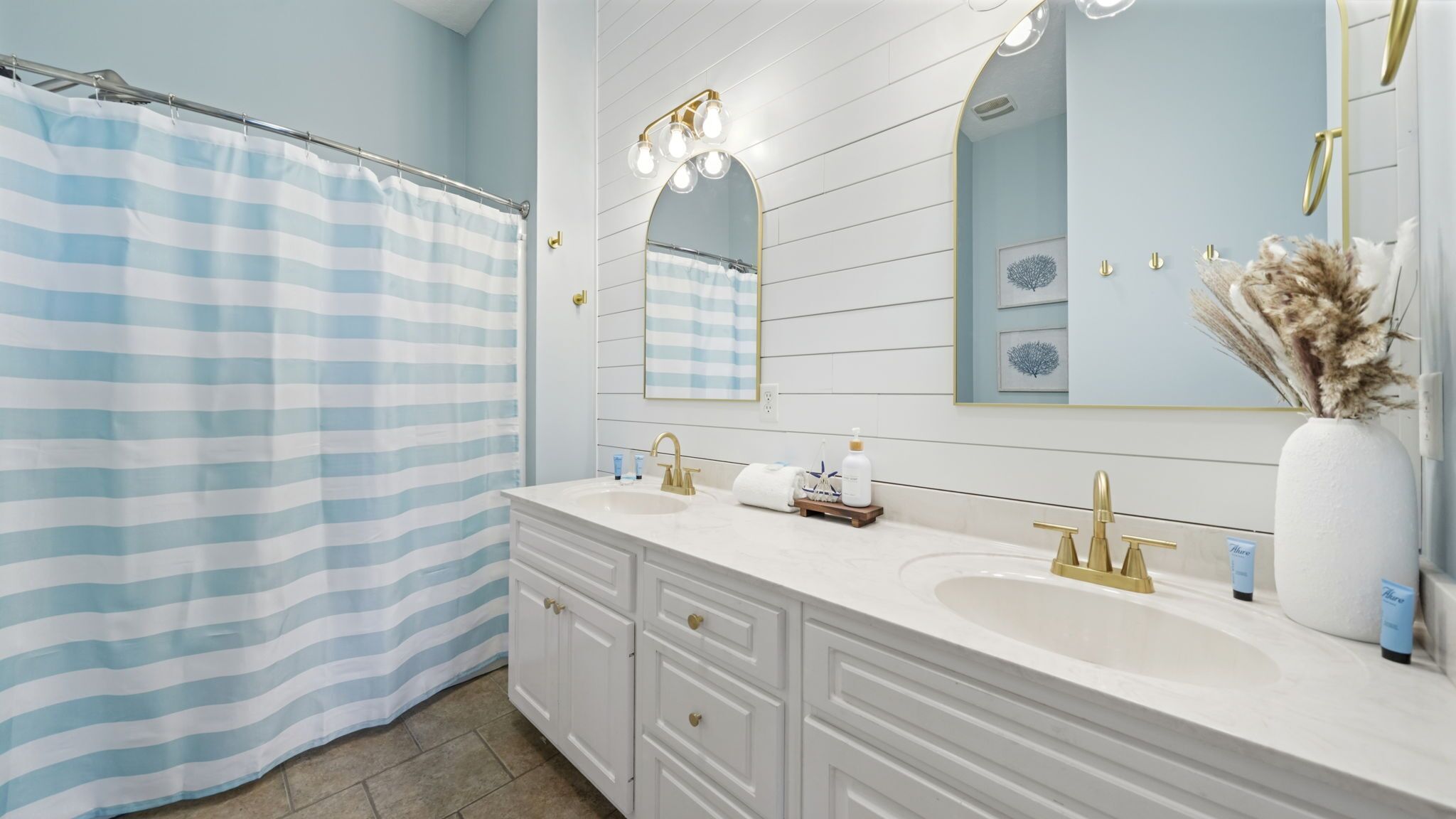 A bathroom with a striped shower curtain, a vanity with two sinks, a mirror, and a decorative vase with dried plants.