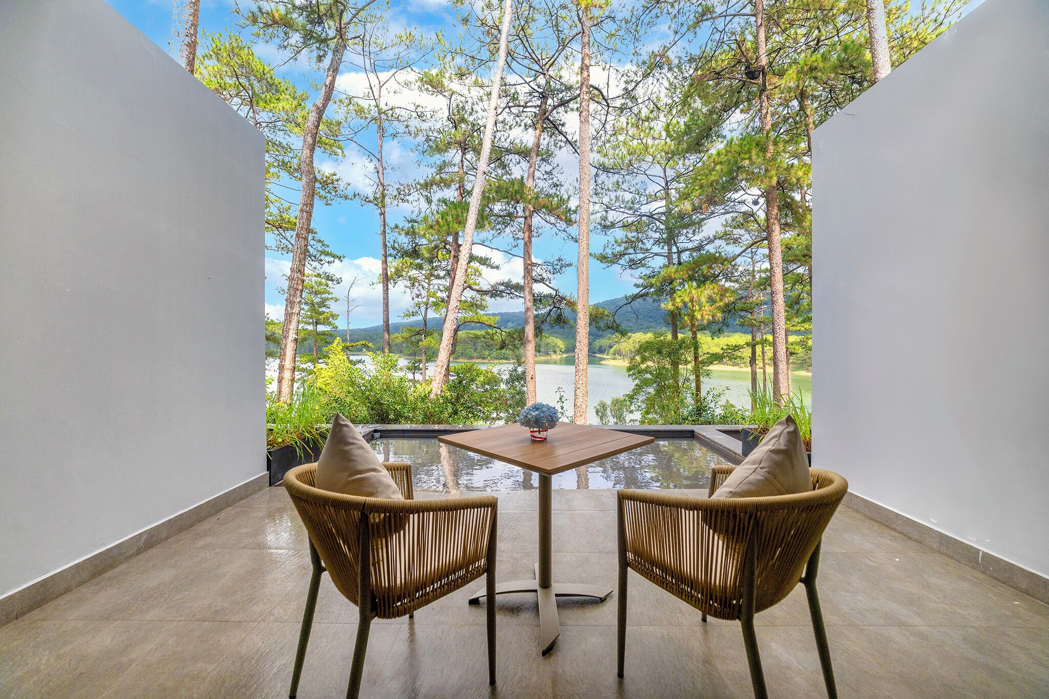 Ein Balkon mit zwei Korbstühlen und einem kleinen Tisch, mit Blick auf eine üppige Landschaft mit Bäumen und einem Gewässer.