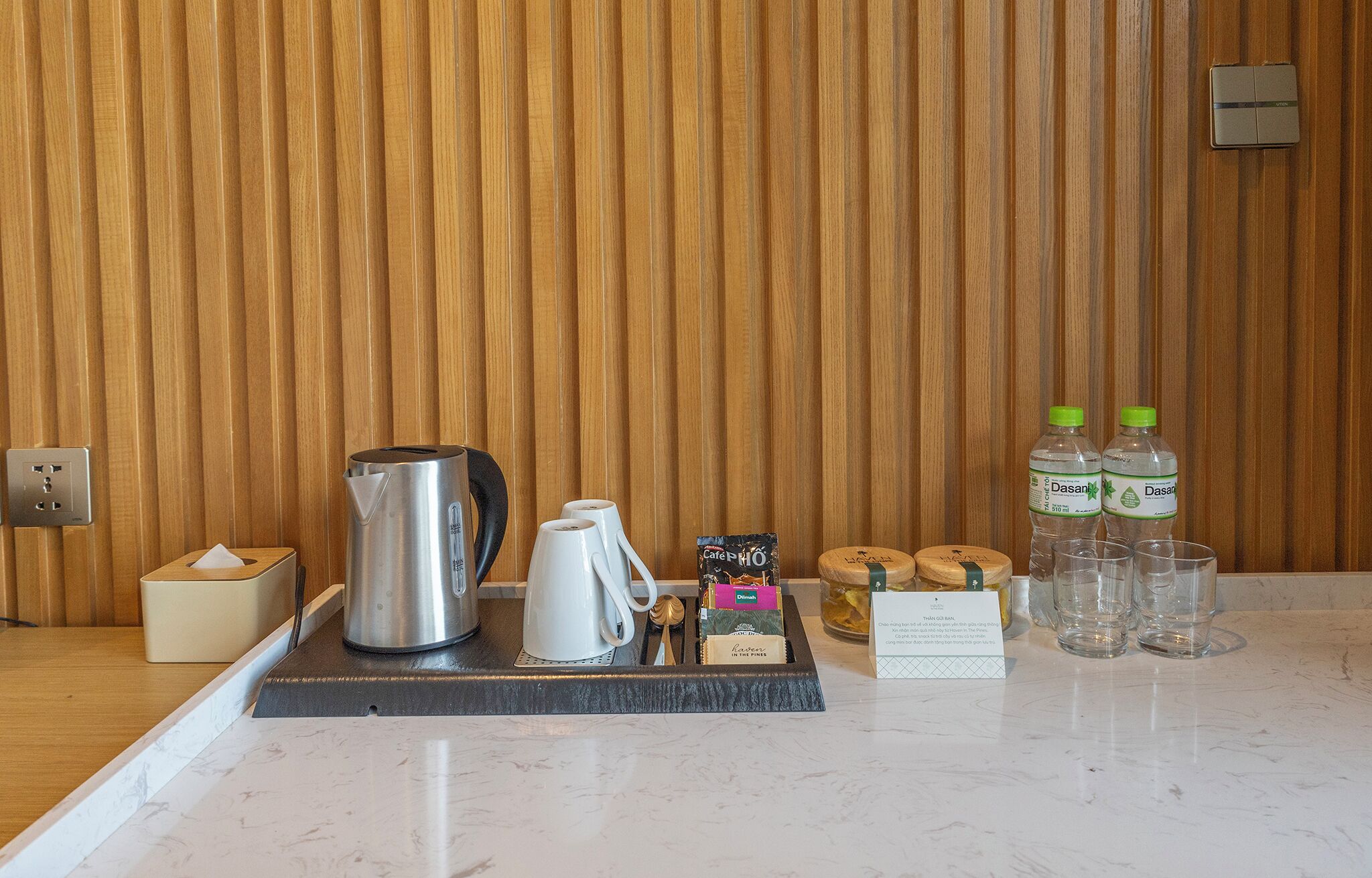 A hotel room's breakfast area with a wooden wall, a coffee maker, a kettle, a tray with coffee cups, a box of coffee pods, Dasani water bottles, and a tissue box.