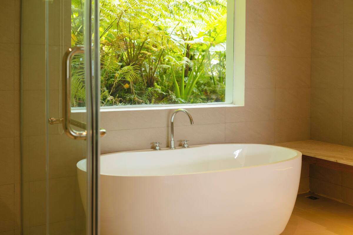 A modern bathroom with a freestanding bathtub, a glass shower enclosure, and a window with a view of greenery.