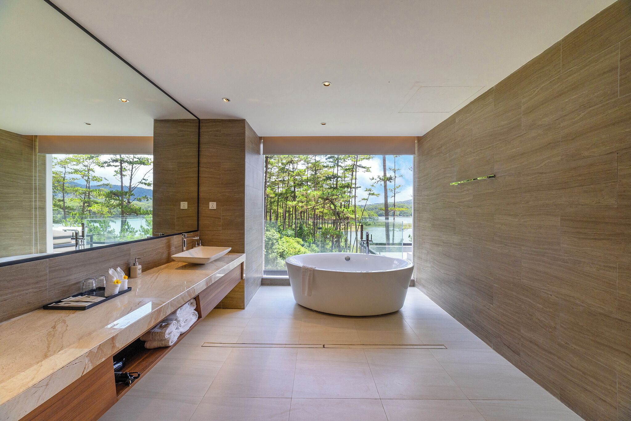 Une salle de bain moderne avec une grande baignoire, un meuble-lavabo avec un miroir et une vue sur les arbres et l’eau.
