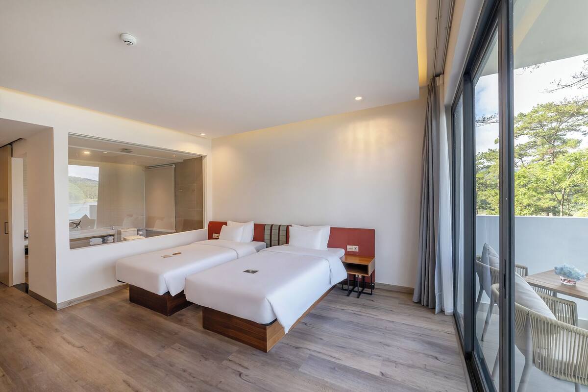 A modern hotel room with two beds, a large mirror, and a view of trees through the window.