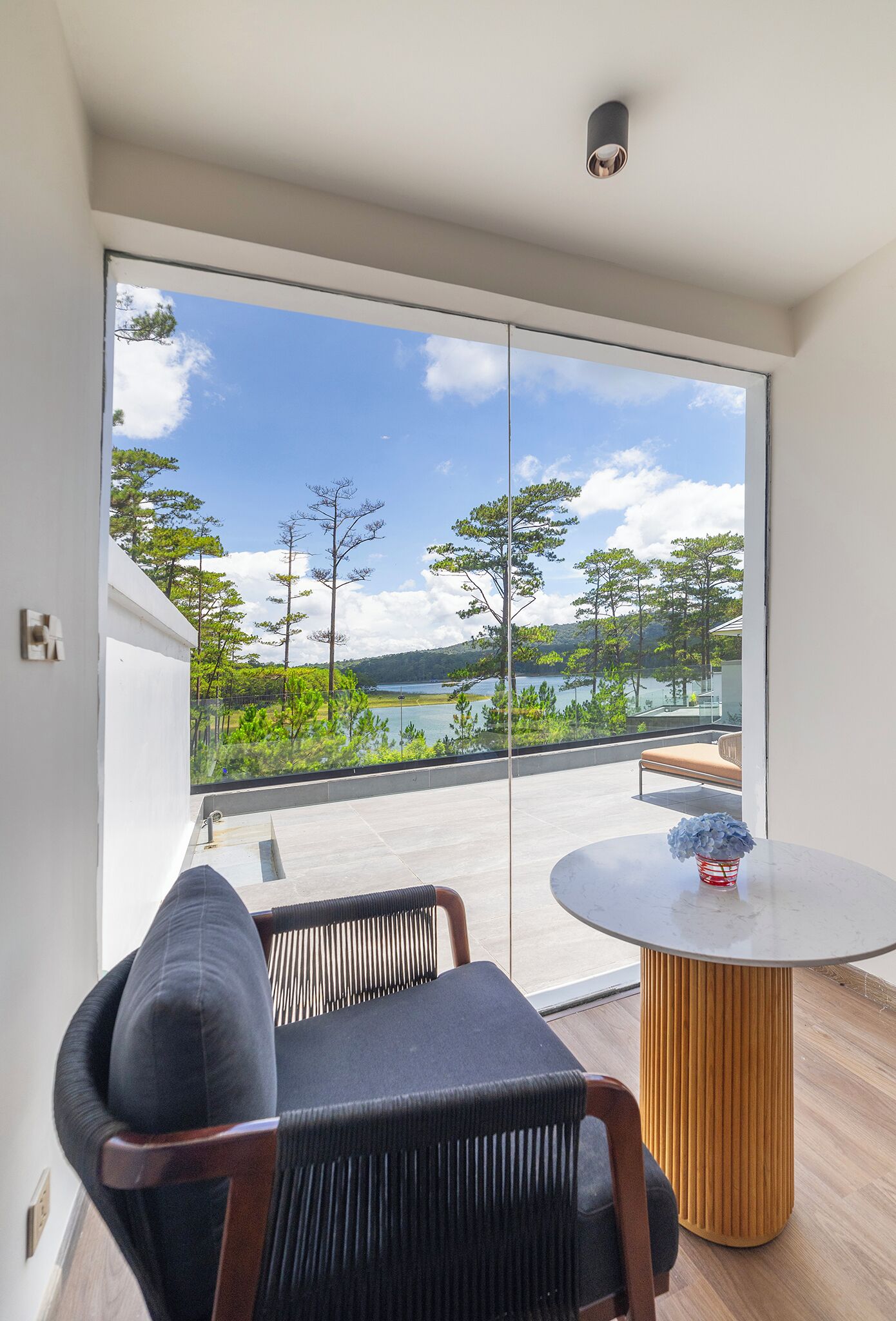 A balcony with a view of a lake and trees, a round table with a vase, and a chair.
