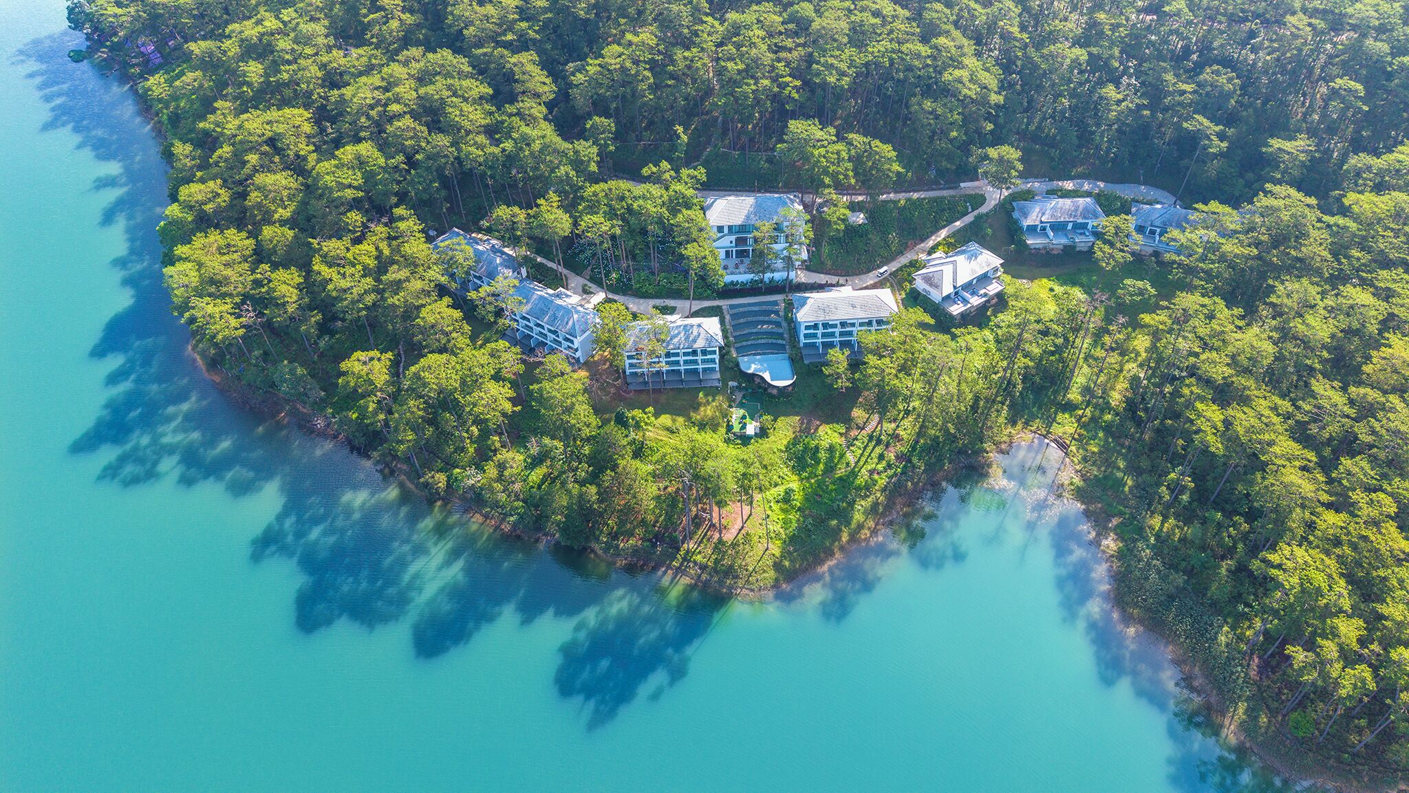 Oversigtsbillede af et resort med flere bygninger omgivet af tæt skov og en klar blå sø.