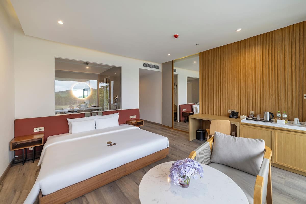 A modern hotel room with a large bed, a round coffee table, a seating area, a kitchenette, and a large mirror.