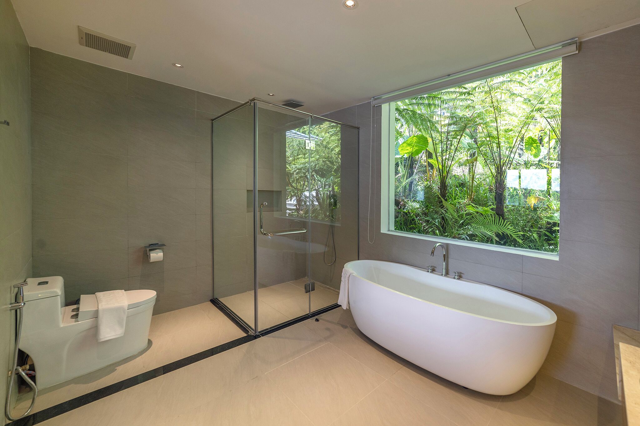 A modern bathroom with a glass-enclosed shower, a freestanding bathtub, and a window providing a view of greenery.