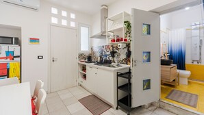 Eine kompakte Küche mit weißen Schränken, einer Mikrowelle und einer Kaffeemaschine. Eine Tür führt zu einem Badezimmer mit Dusche und Toilette.