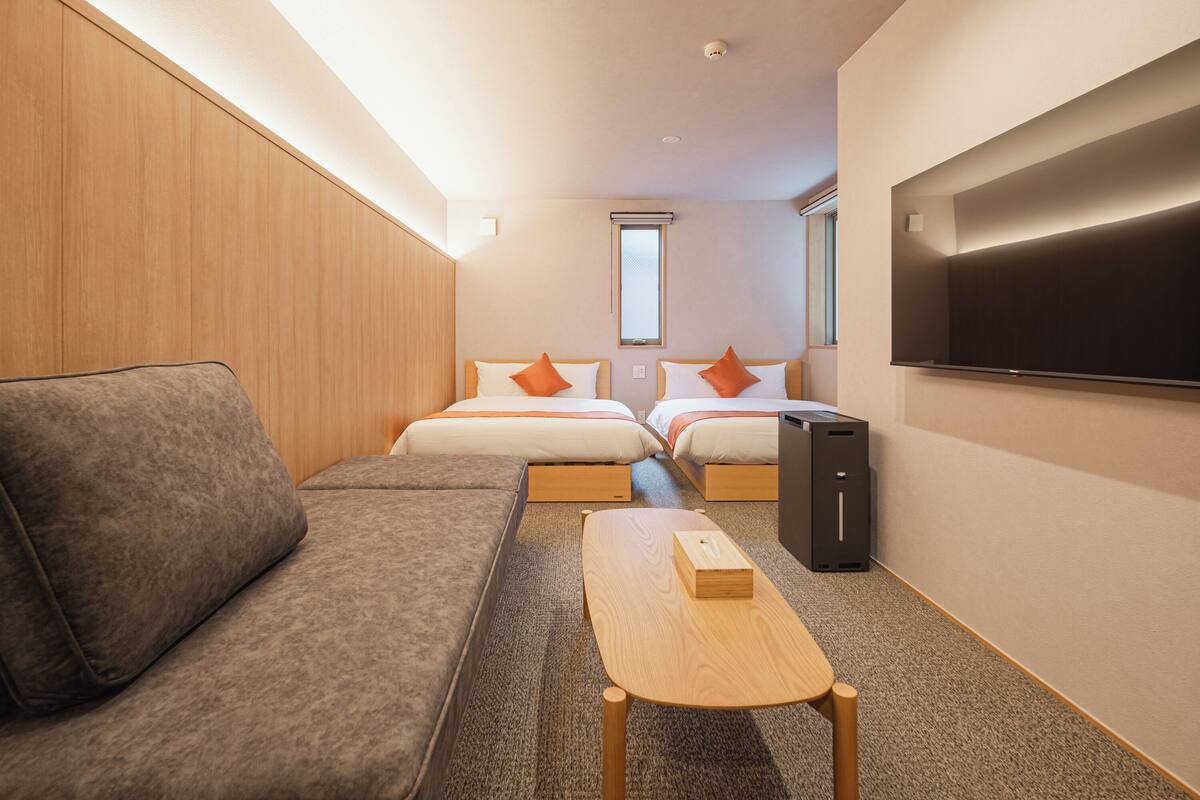 A compact hotel room with a sofa, two single beds with orange pillows, a TV, and a desk with a box.